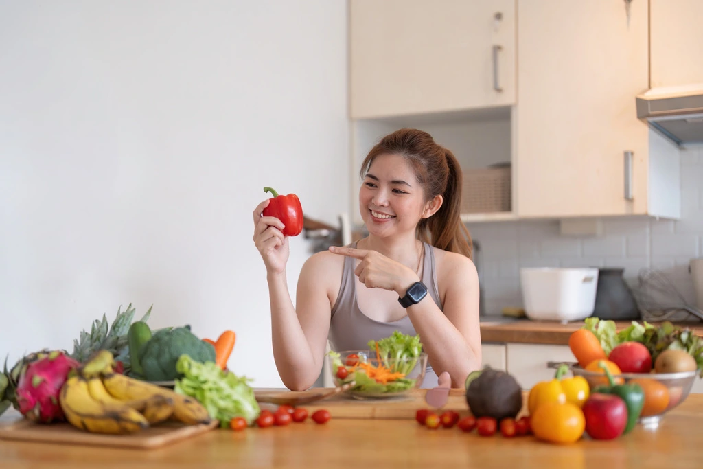 Jess Carter of CravyBite Kitchen smiling as she points to a fresh red bell pepper, with a variety of colorful fruits and vegetables on her kitchen counter.