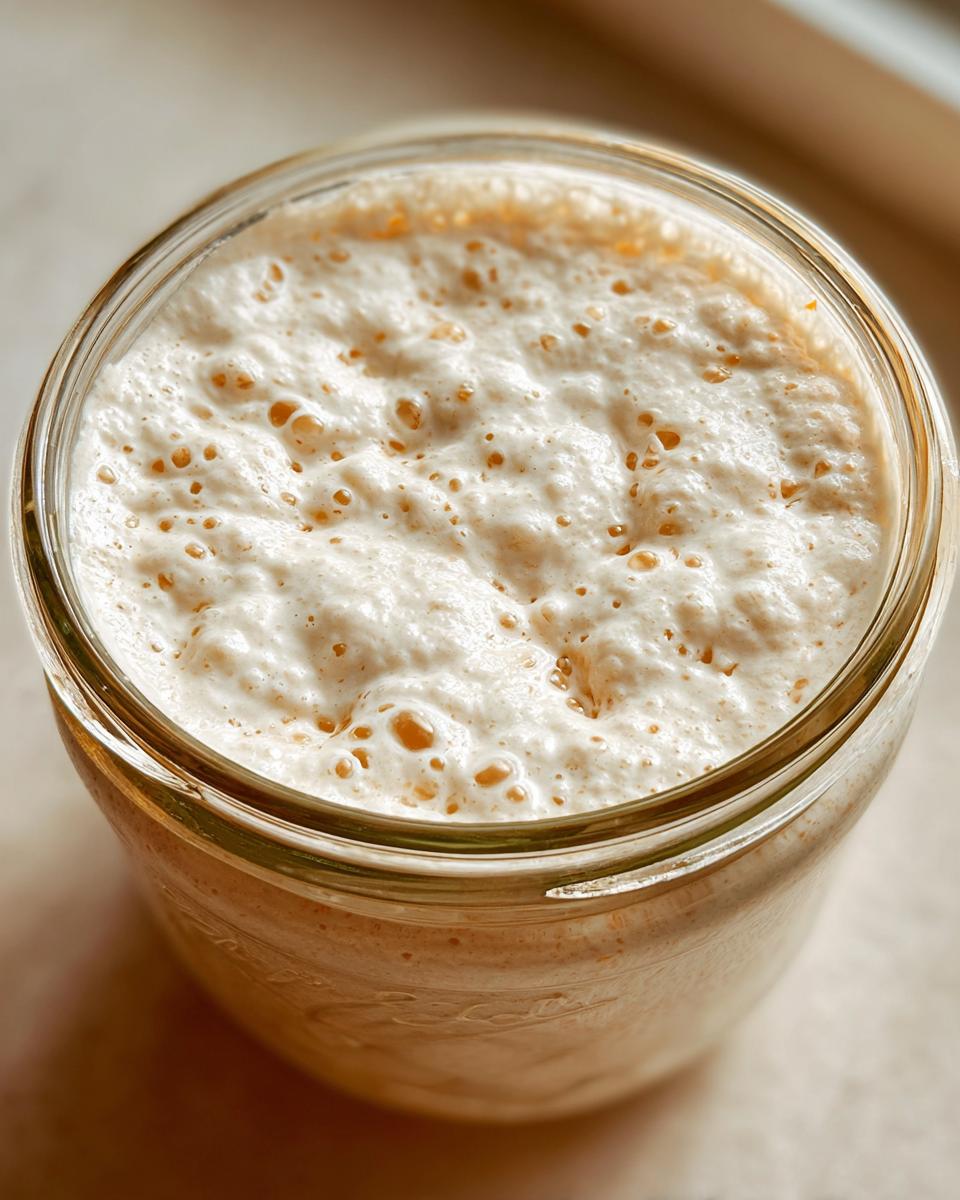 Overhead view of a very active sourdough starter in a glass jar, showing a thick layer of foam and bubbles.