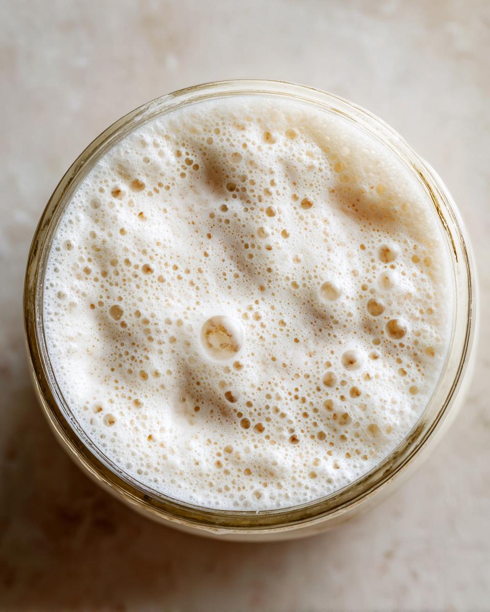 Overhead view of a highly active sourdough starter, showing a thick layer of foam and bubbles in a glass jar.