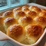 A close-up of twelve golden brown, glossy dinner rolls baked together in a white dish, brushed with butter.