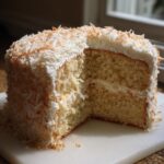 Cross-section of a moist coconut cake showing layers of white cake and frosting, topped with toasted coconut flakes.
