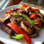 Close-up of perfectly cooked steak fajitas featuring sliced steak, vibrant red and green bell peppers, and caramelized onions on a white plate.