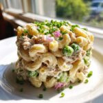 A cylindrical serving of creamy tuna pasta salad featuring elbow macaroni, green peas, red onion, and topped with fresh chives.