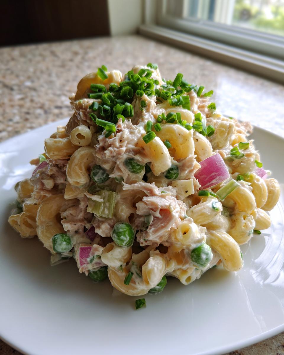 A scoop of creamy tuna pasta salad featuring elbow macaroni, peas, red onion, and topped with fresh chives.