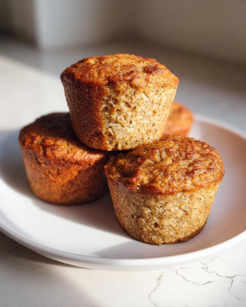A stack of three golden-brown banana protein muffins resting on a white plate, catching bright sunlight.