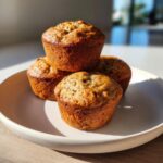 Four golden-brown banana protein muffins stacked on a light-colored plate, beautifully lit by sunlight.