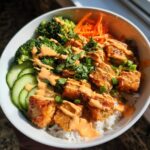 A close-up of a white bowl filled with Bang Bang Salmon Bowls featuring crispy salmon pieces over rice, topped with orange sauce, broccoli, carrots, and cucumbers.