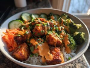 A close-up of a bowl featuring crispy salmon pieces drizzled with orange sauce, served over rice with broccoli, shredded carrots, and cucumber slices for a Bang bang salmon bowls.