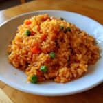 A close-up of a white bowl filled with bright orange Spanish rice mixed with green peas and diced carrots.