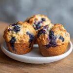 Three freshly baked blueberry protein muffins with golden tops, served on a speckled white plate.