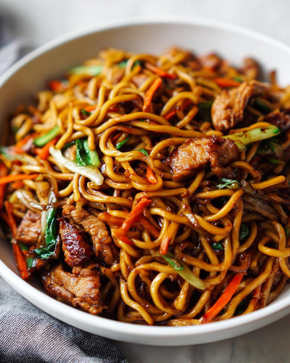 Close-up of a white bowl filled with glossy chicken chow mein recipe noodles, chicken pieces, and colorful vegetables.