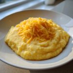 A close-up of creamy cheese grits served in a white bowl, generously topped with shredded cheddar cheese.