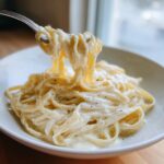 A fork lifting creamy fettuccine noodles coated in rich olive garden alfredo sauce, seasoned with black pepper.