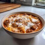 A close-up of a bowl filled with rich, red creamy lasagna soup, topped with dollops of white ricotta and grated cheese.