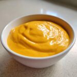 A close-up of a small white bowl filled with thick, bright orange homemade nacho cheese sauce.