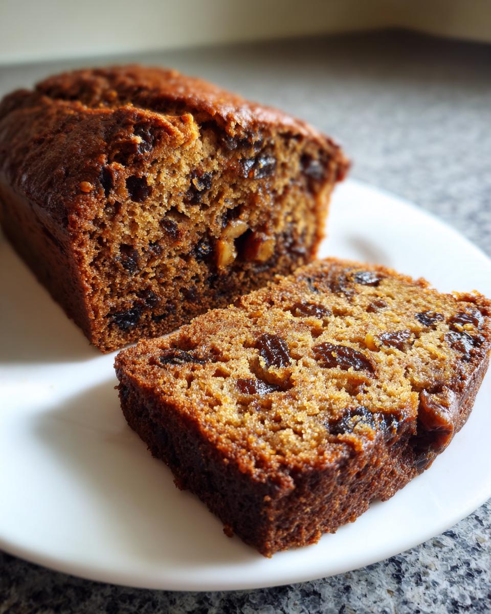 Close-up of a freshly baked loaf of date nut bread with one thick slice cut and resting beside it on a white plate.