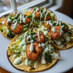 Four grilled shrimp taco servings on corn tortillas, topped with shredded lettuce and a drizzle of light green sauce.