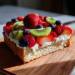 A close-up of a slice of fruit pizza sugar cookie topped with cream cheese frosting, strawberries, blueberries, and kiwi.