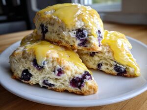 Close-up of three delicious lemon blueberry scones stacked on a white plate, generously topped with bright yellow lemon glaze.