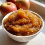 A small white bowl filled with chunky, spiced applesauce, with fresh red apples blurred in the background.