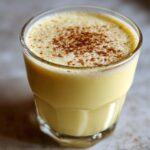 Close-up of a glass filled with creamy, yellow homemade eggnog, topped with a dusting of cinnamon.
