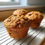 Three golden brown honey bran muffins topped with raisins cooling on a wire rack near a window.