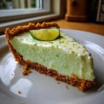 A close-up of a creamy slice of key lime cheesecake with a graham cracker crust, topped with lime zest and a lime wheel.