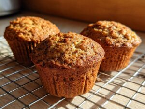 Three freshly baked, golden brown bran muffins resting on a wire cooling rack in bright sunlight.