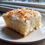 A moist slice of coconut poke cake topped with white frosting and toasted coconut flakes.
