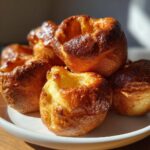 Close-up of several golden brown, perfectly risen yorkshire pudding cups stacked on a white plate.