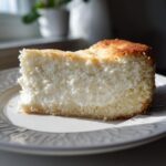 A thick slice of creamy, baked Protein Cheesecake with a golden-brown top, served on a decorative white plate.