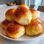 A stack of freshly baked, glossy sourdough discard rolls with a golden-brown crust on a white plate.