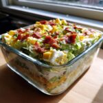 A clear glass dish showcasing the distinct layers of an amazing 7 layer salad, topped with bacon and egg.