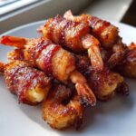 Close-up of several perfectly cooked bacon wrapped shrimp piled on a white plate, showing a caramelized glaze.