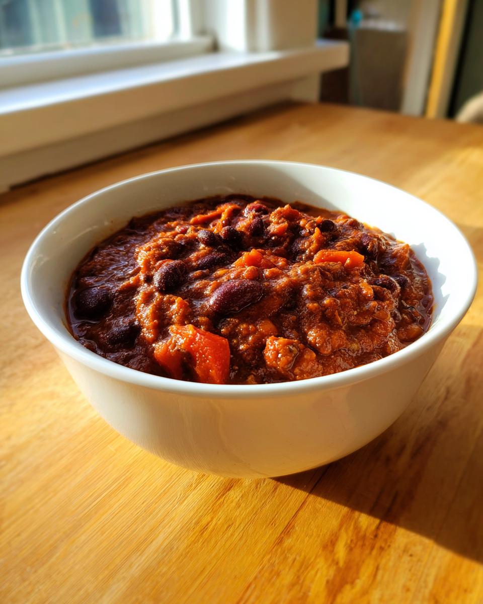 A white bowl filled with thick, rich black bean chili, set on a sunlit wooden table near a window.