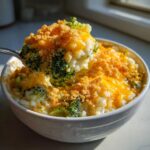A spoonful of creamy, cheesy broccoli rice is lifted from a white bowl, topped with melted cheddar and crispy breadcrumbs.