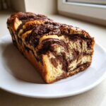 A freshly baked, golden-brown chocolate babka loaf showing rich, dark chocolate swirls inside, resting on a white plate.