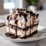 A decadent slice of layered ice cream cake topped with whipped cream, chocolate drizzle, and cookie crumbs.