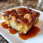 A close-up of a moist slice of bread pudding with bourbon sauce generously drizzled over the top and pooling on the white plate.