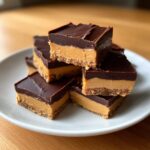 A stack of rich buckeye fudge squares featuring a peanut butter layer and a thick chocolate topping on a white plate.