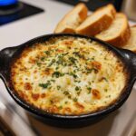 Close-up of a hot, cheesy baked bread dip topped with parsley, served with slices of toasted bread.