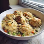Close-up of a bowl filled with creamy chicken and dumplings with biscuits, peas, and carrots.