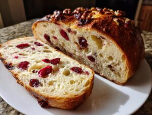 A loaf of homemade cranberry walnut bread, sliced to show the soft interior filled with red cranberries and topped with walnuts.
