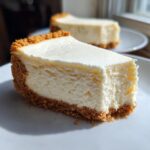 Close-up of a thick slice of creamy cheesecake recipe with a golden graham cracker crust on a white plate.