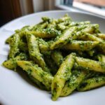 A close-up shot of penne pasta coated generously in vibrant green creamy pesto pasta sauce, served in a white bowl.