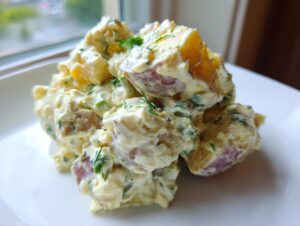 A close-up of a serving of creamy red potato salad mixed with herbs on a white plate.