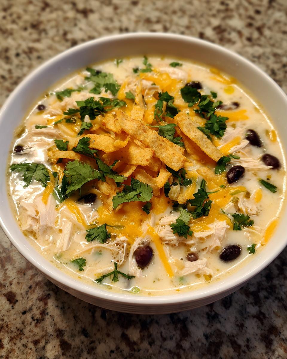 A close-up of a bowl of creamy tortilla soup topped with shredded chicken, black beans, cheese, cilantro, and tortilla strips.