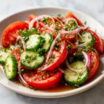 Close-up of a fresh cucumber tomato salad featuring sliced tomatoes, cucumbers, red onion, and fresh herbs in a light dressing.