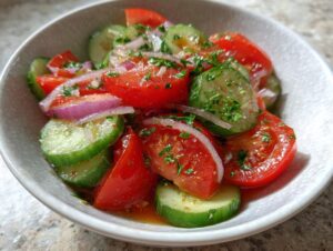 Close-up of a vibrant cucumber tomato salad featuring sliced cucumbers, wedges of red tomato, and slivers of red onion, topped with herbs.