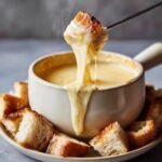 A piece of toasted bread is dipped into a steaming pot of creamy cheese fondue recipe, dripping with melted cheese.